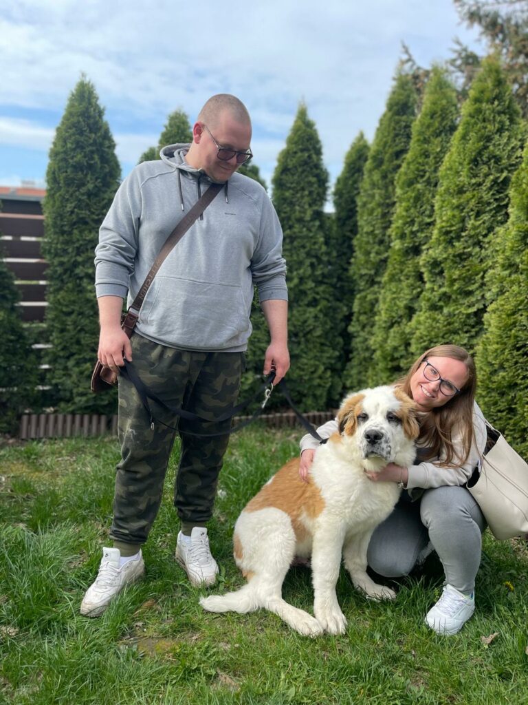 Na zdjęciu widać kobietę i mężczyznę stojących na trawie obok dużego, biało-brązowego psa na smyczy. Mężczyzna trzyma smycz, a kobieta kuca obok psa i go przytula. W tle znajdują się wysokie, zielone krzewy oraz ogrodzenie.