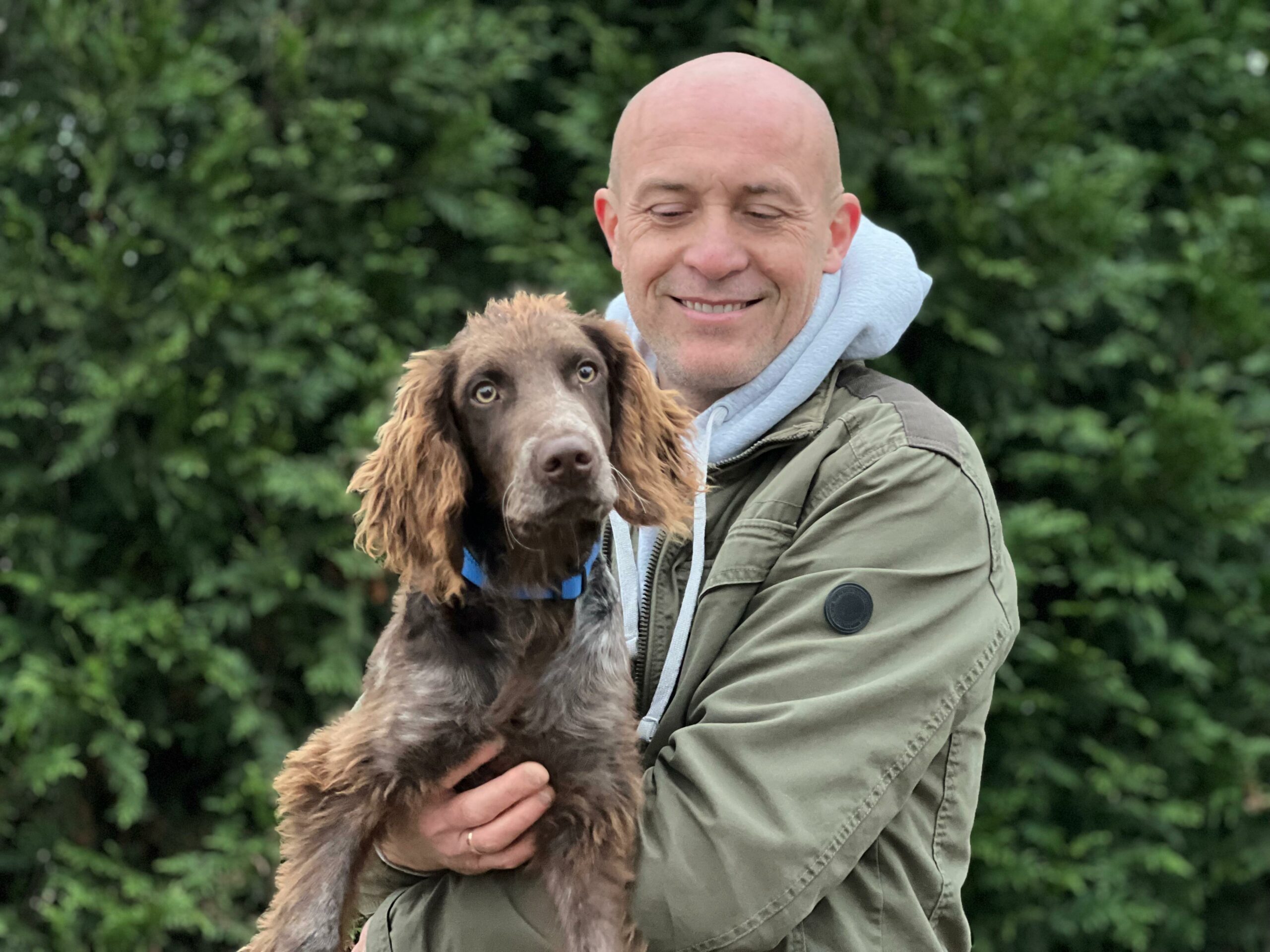 Na zdjęciu widać mężczyznę stojącego na zewnątrz, na tle zielonych krzewów. Trzyma na rękach brązowego psa o dłuższej, falującej sierści i niebieskiej obroży. Mężczyzna ma na sobie zieloną kurtkę z kapturem i lekko się uśmiecha, a pies patrzy prosto w stronę aparatu.