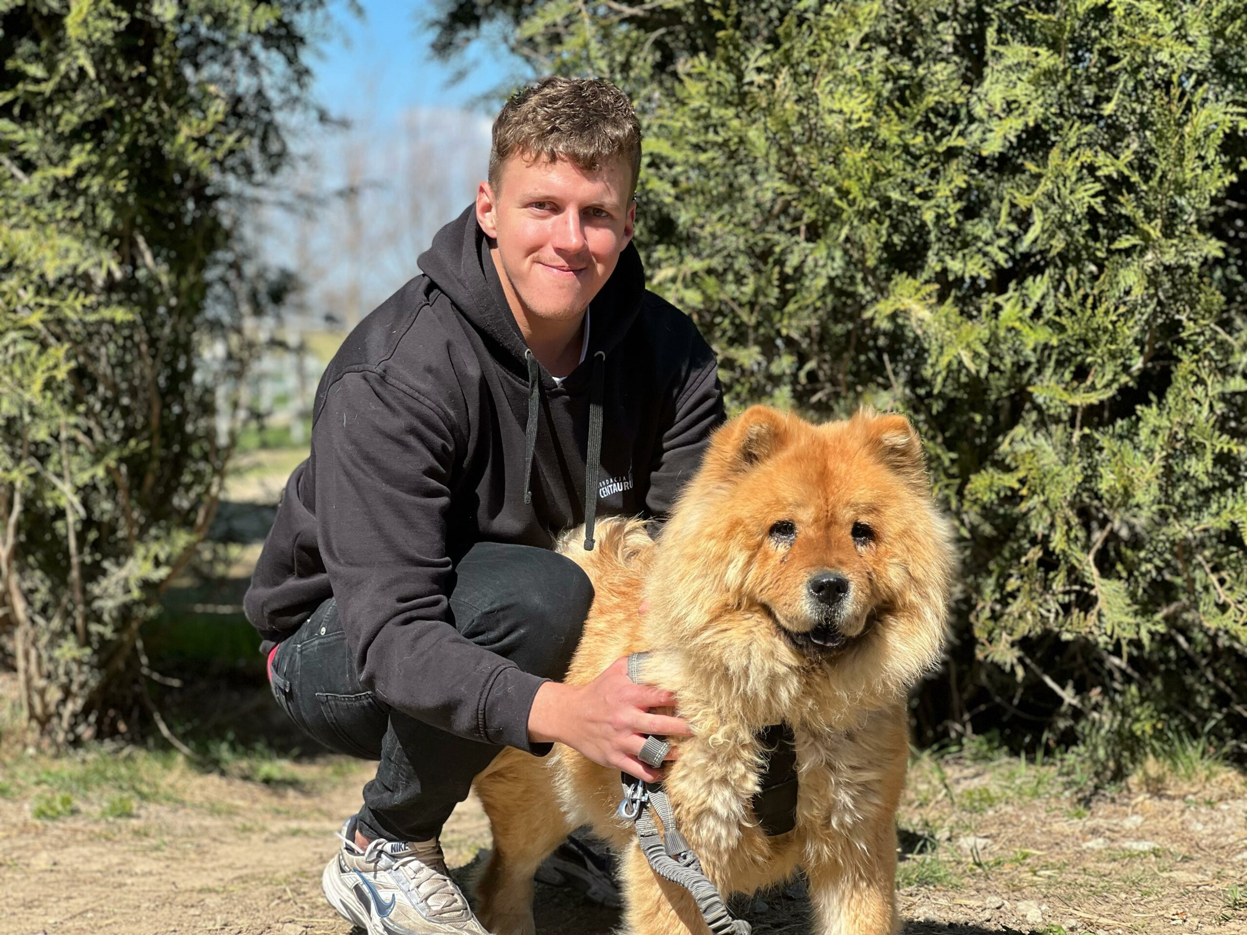 Na zdjęciu widać młodego mężczyznę kucającego obok dużego, puszystego psa o rudobrązowej sierści. Mężczyzna trzyma psa za smycz i delikatnie go obejmuje. Znajdują się na zewnątrz, w słoneczny dzień, a w tle widoczne są wysokie, zielone krzewy.