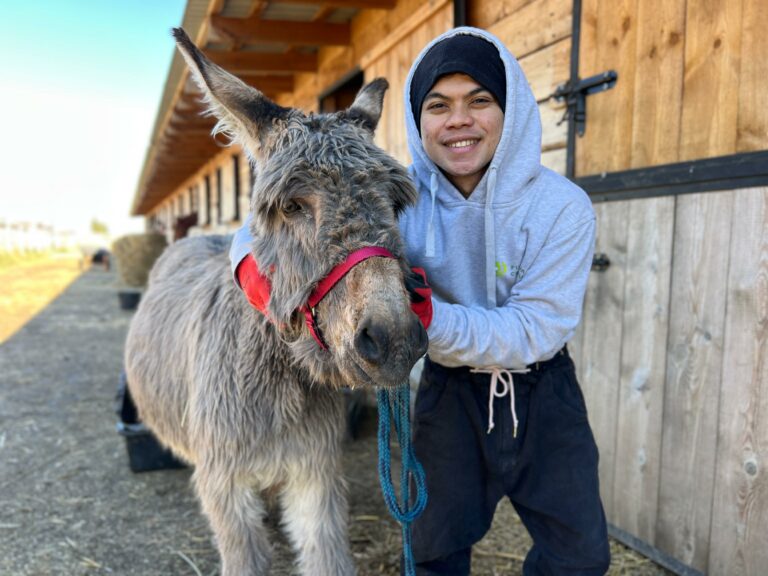 Na zdjęciu widzimy osobę w szarej bluzie z kapturem, czarnych spodniach i czarnej czapce, która z uśmiechem obejmuje głowę osła. Zwierzę ma kudłatą sierść i założony czerwono-szary kantar. Tło stanowi drewniana stajnia z otwartymi drzwiami, a na ziemi widać słomę.