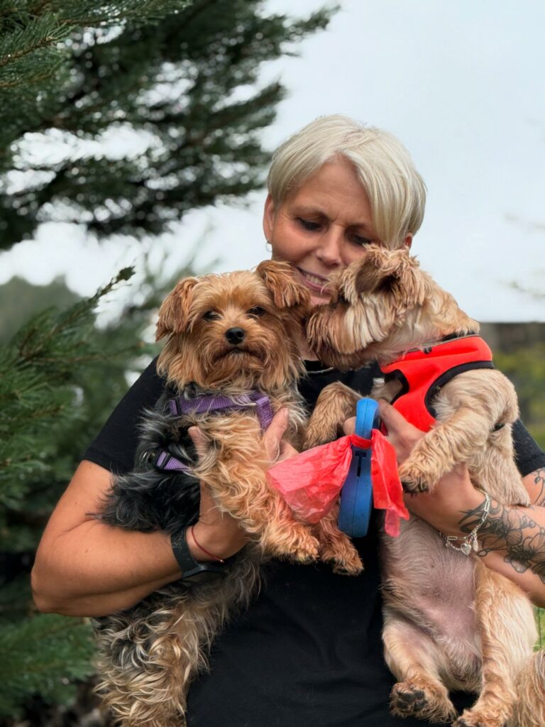 Na zdjęciu widać kobietę trzymającą w ramionach dwa małe psy. Jeden z nich ma fioletową uprząż, a drugi czerwoną kamizelkę i przypiętą smycz. Kobieta stoi na zewnątrz, a w tle widoczna jest zieleń drzew. Psy są przytulone do niej, a ona patrzy na nie z czułością.