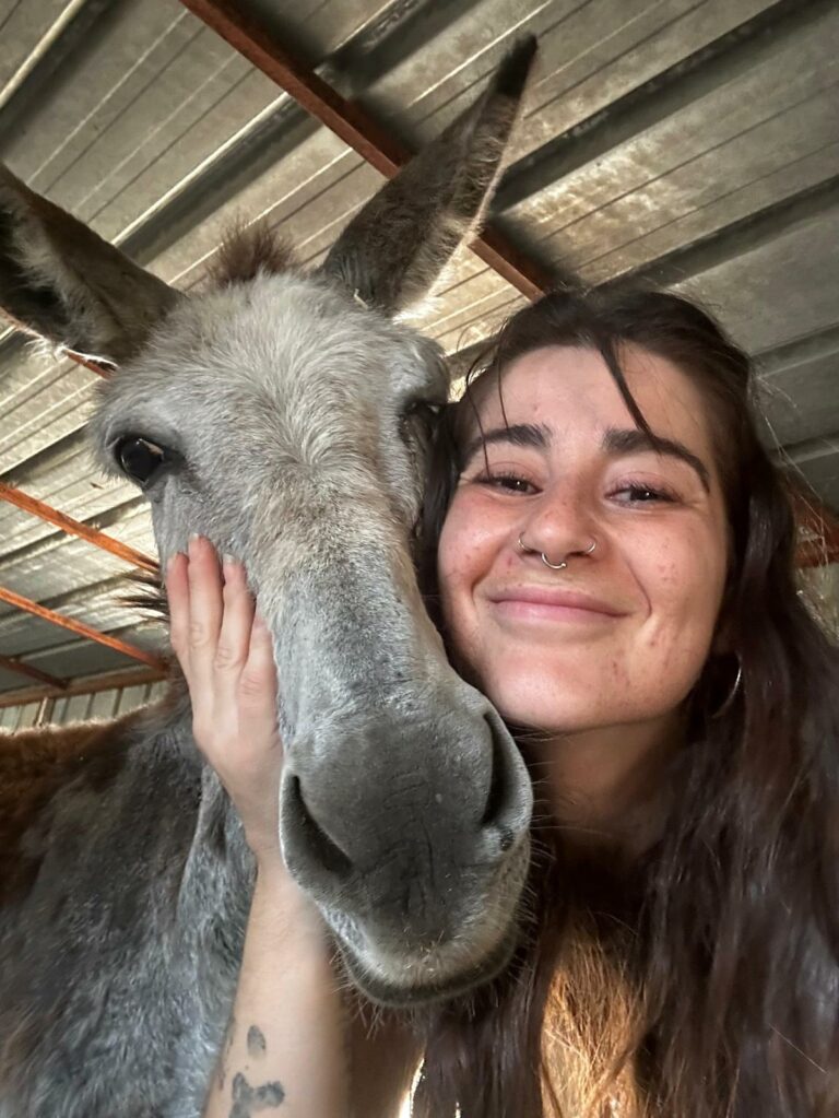 Na zdjęciu widzimy zbliżenie uśmiechniętej kobiety przytulającej się do osła w metalowej wiacie. Twarz kobiety jest delikatnie oparta o pysk zwierzęcia, a jedna ręka obejmuje jego policzek z czułością. Osioł ma duże uszy i spokojne spojrzenie, a kobieta — długie ciemne włosy, piercing w nosie i widoczne tatuaże na ramieniu.