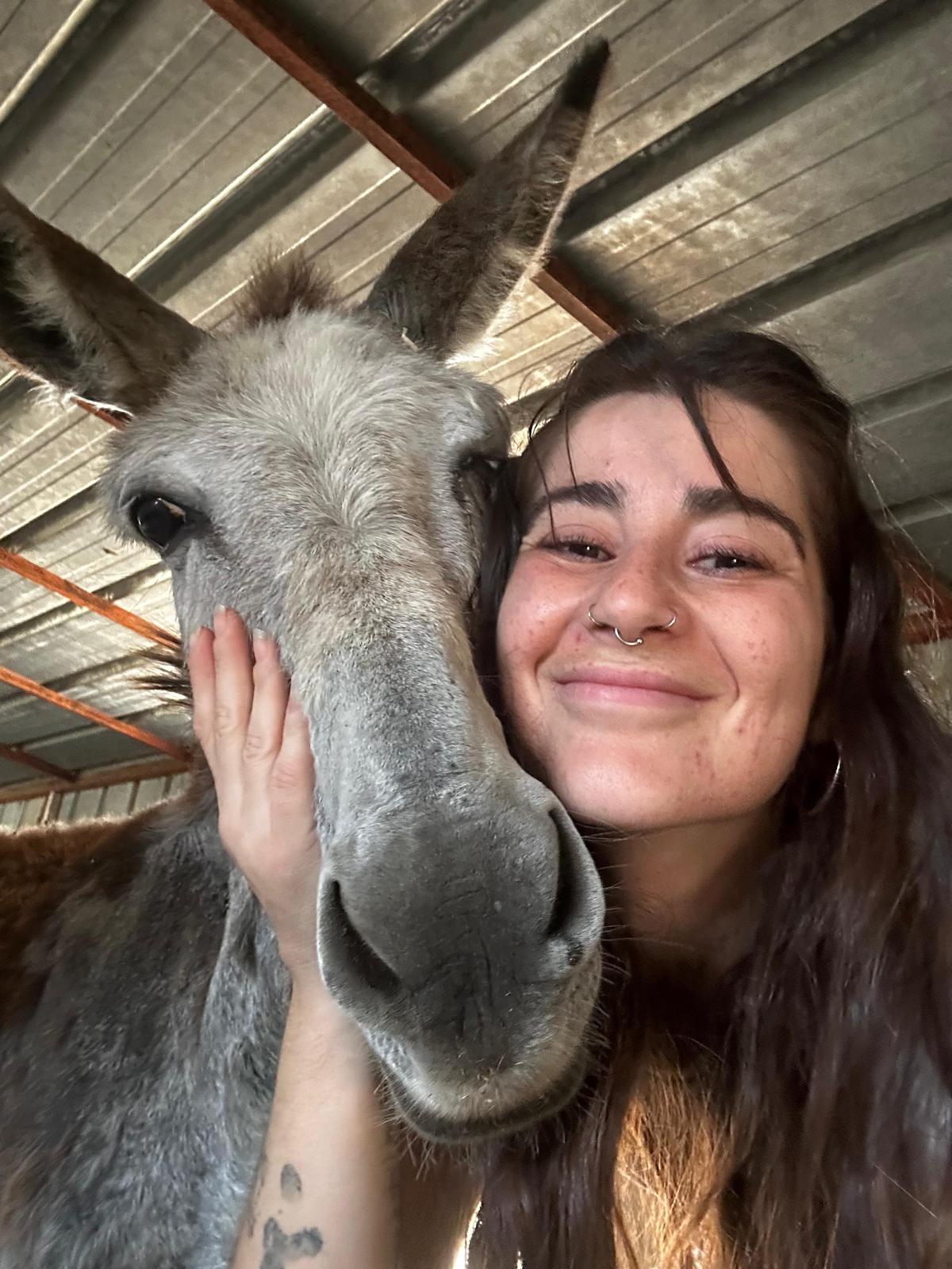 Na zdjęciu widzimy zbliżenie uśmiechniętej kobiety przytulającej się do osła w metalowej wiacie. Twarz kobiety jest delikatnie oparta o pysk zwierzęcia, a jedna ręka obejmuje jego policzek z czułością. Osioł ma duże uszy i spokojne spojrzenie, a kobieta — długie ciemne włosy, piercing w nosie i widoczne tatuaże na ramieniu.