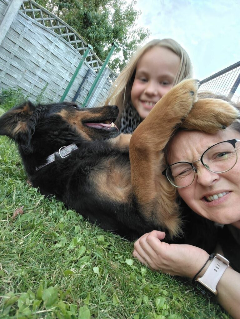 Na zdjęciu widać kobietę i dziewczynkę leżące na trawie obok czarno-brązowego psa. Pies przewraca się na bok i opiera łapę na twarzy kobiety. Wszyscy wyglądają na rozbawionych i bawią się na zewnątrz.