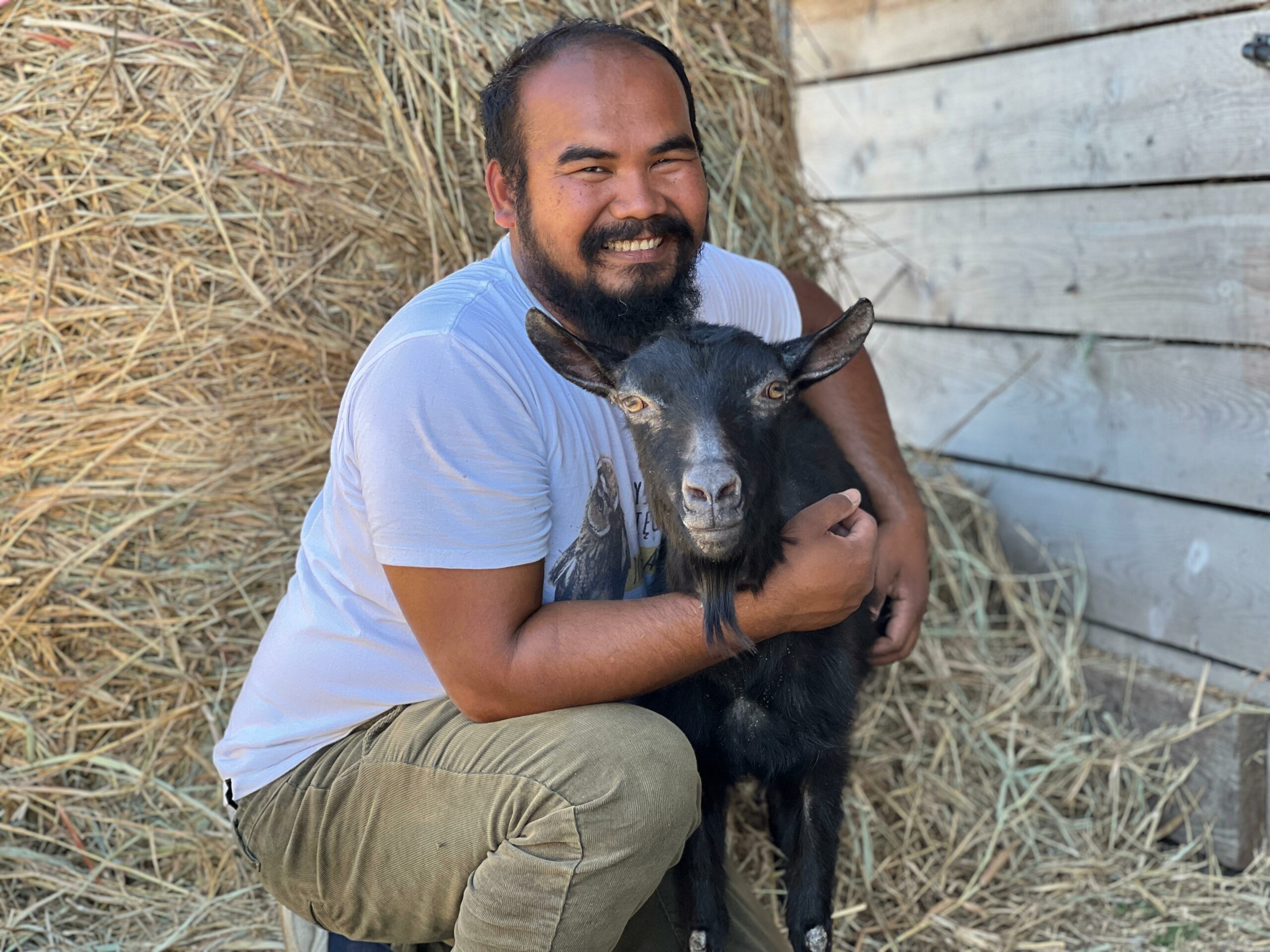 Na zdjęciu widzimy mężczyznę w białej koszulce i khaki spodniach, który kuca i uśmiecha się, trzymając czarną kozę. Zwierzę ma krótką sierść z jaśniejszymi akcentami na pysku i nogach, a jego spokojna postawa sugeruje zaufanie do człowieka. Tło tworzą bele siana i drewniane deski.