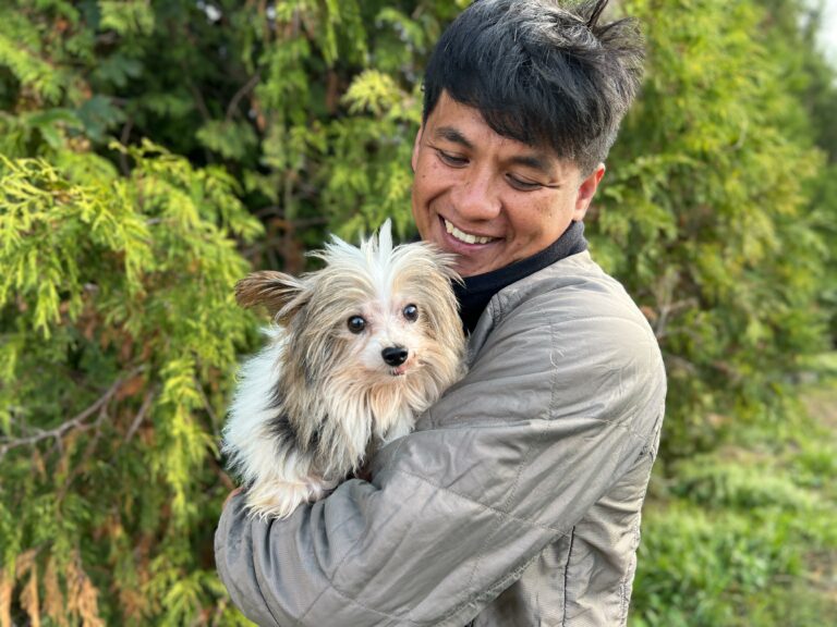 Na zdjęciu widać uśmiechniętego mężczyznę trzymającego na rękach małego, puszystego psa o jasnej sierści. Stoją na zewnątrz, a w tle widoczna jest zielona roślinność. Mężczyzna patrzy na psa z radością, a pies skierowany jest w stronę aparatu.