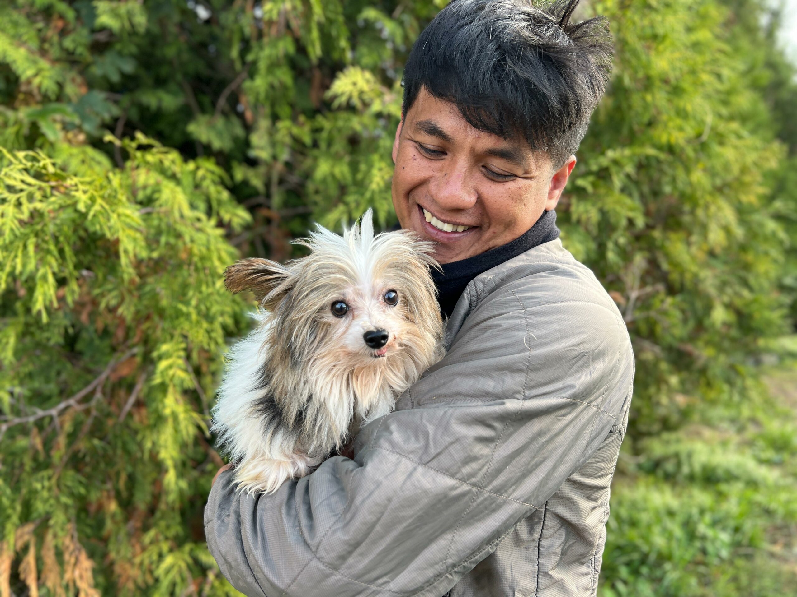 Na zdjęciu widać uśmiechniętego mężczyznę trzymającego na rękach małego, puszystego psa o jasnej sierści. Stoją na zewnątrz, a w tle widoczna jest zielona roślinność. Mężczyzna patrzy na psa z radością, a pies skierowany jest w stronę aparatu.