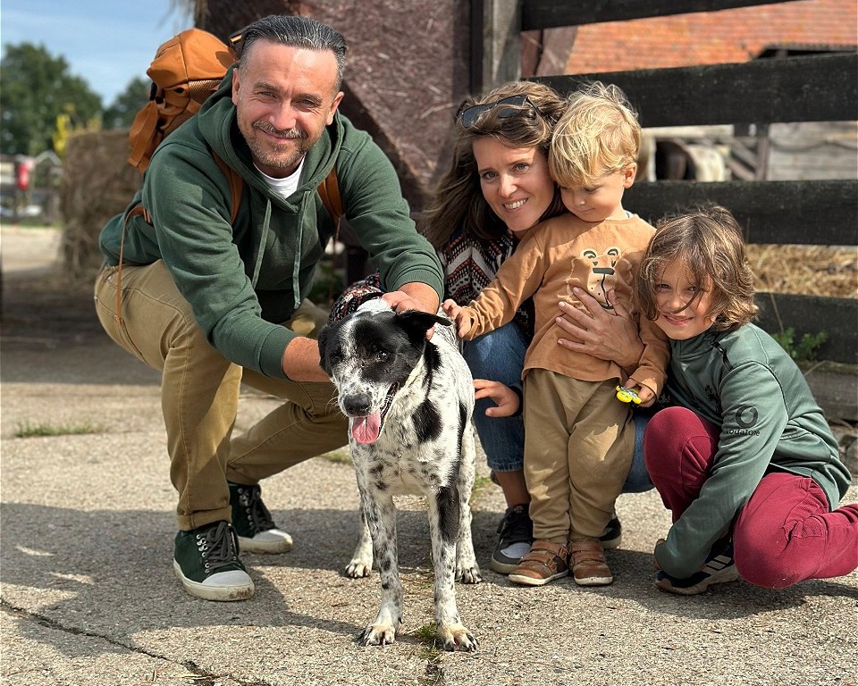 Na zdjęciu widać rodzinę – mężczyznę, kobietę oraz dwoje dzieci – stojących na zewnątrz obok czarno-białego psa. Wszyscy są uśmiechnięci i dotykają psa, który stoi przed nimi z otwartym pyskiem. Tło sugeruje wiejskie lub podmiejskie otoczenie z budynkiem i ogrodzeniem.