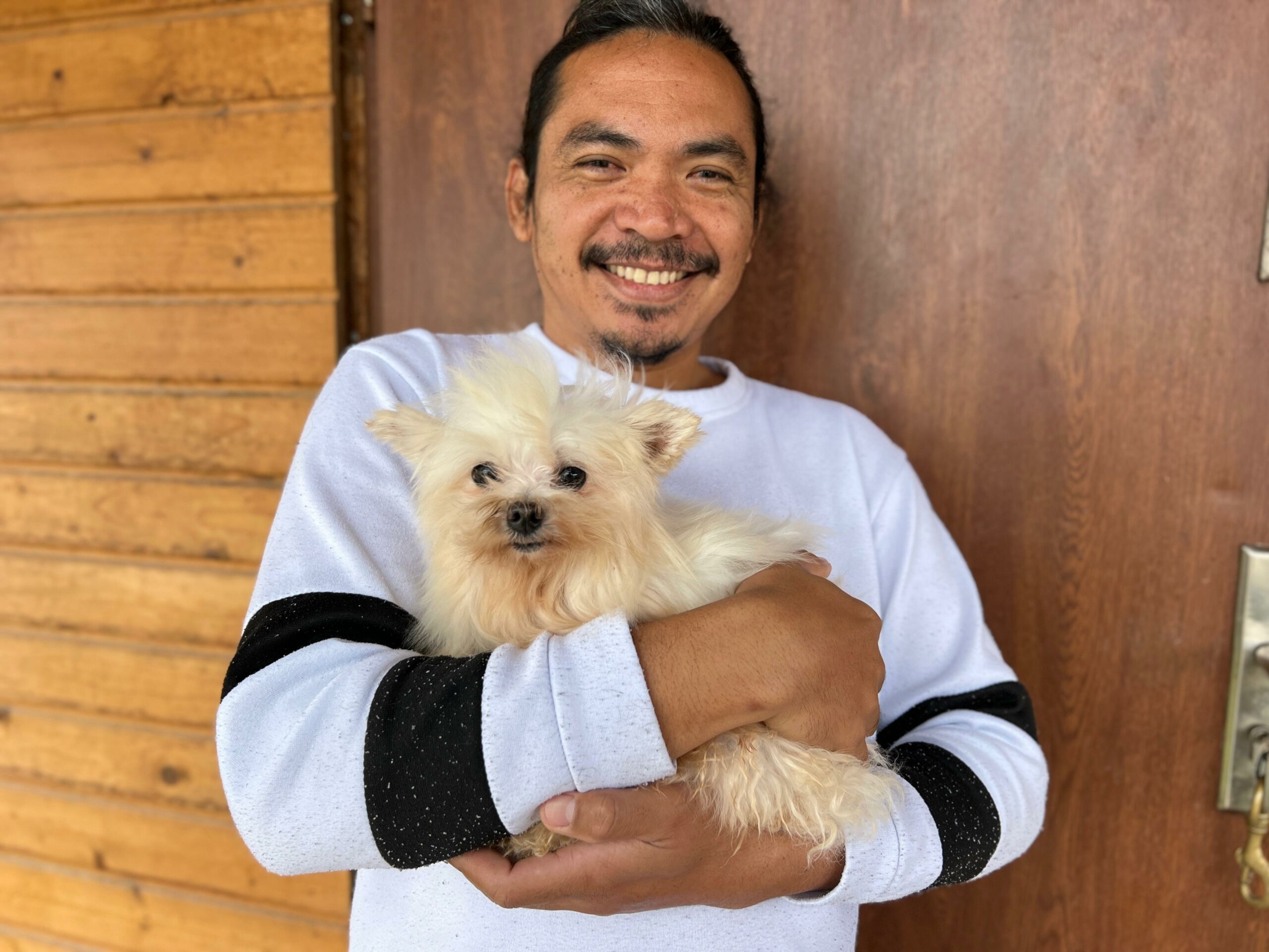 Na zdjęciu widać uśmiechniętego mężczyznę stojącego przed drewnianą ścianą i drzwiami. Trzyma na rękach małego, jasnego psa o puszystej sierści. Mężczyzna patrzy w stronę aparatu, a pies ma spokojny wyraz pyska i również jest skierowany w stronę obiektywu.