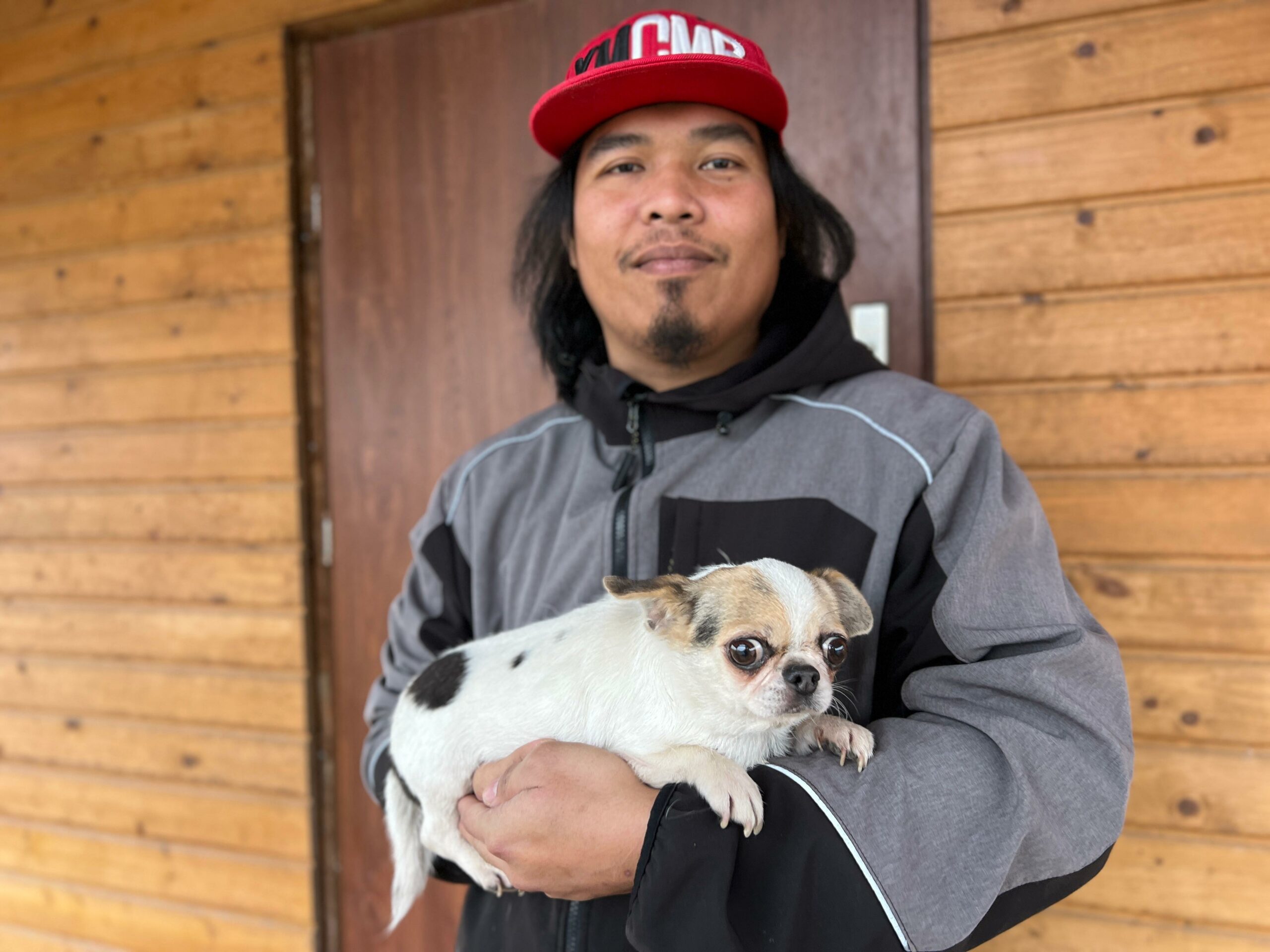Na zdjęciu widać mężczyznę stojącego przed drewnianą ścianą budynku. Ma na sobie szaro-czarną kurtkę i czerwoną czapkę z daszkiem. W ramionach trzyma małego, biało-brązowego psa. Mężczyzna patrzy w stronę aparatu z lekkim uśmiechem, a pies ma szeroko otwarte oczy i skierowany wzrok na bok.