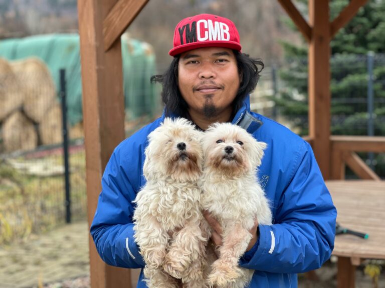 Na zdjęciu widać mężczyznę stojącego na zewnątrz pod drewnianą konstrukcją. Ma na sobie niebieską kurtkę oraz czerwoną czapkę z daszkiem. W rękach trzyma dwa małe, jasne psy o puszystej sierści. W tle widać ogrodzony teren i elementy krajobrazu na świeżym powietrzu.