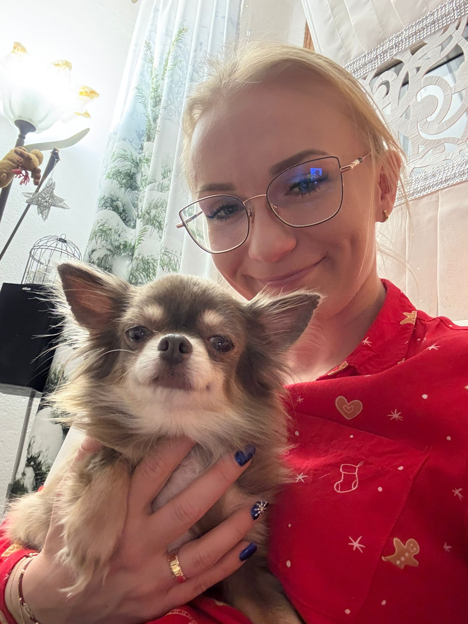 Na zdjęciu widać kobietę w okularach, ubraną w czerwoną koszulę w świąteczne wzory. Trzyma na rękach małego, długowłosego psa o brązowo-białej sierści. Kobieta delikatnie się uśmiecha, a pies patrzy w stronę aparatu. W tle widoczne są zasłony, lampa oraz dekoracje we wnętrzu mieszkania.