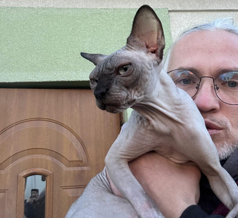 Na zdjęciu widzimy osobę trzymającą kota rasy sfinks, znanego z braku sierści i pomarszczonej skóry. Człowiek ma jasne włosy, okulary i stoi na tle drewnianych drzwi oraz zielono-beżowej ściany.